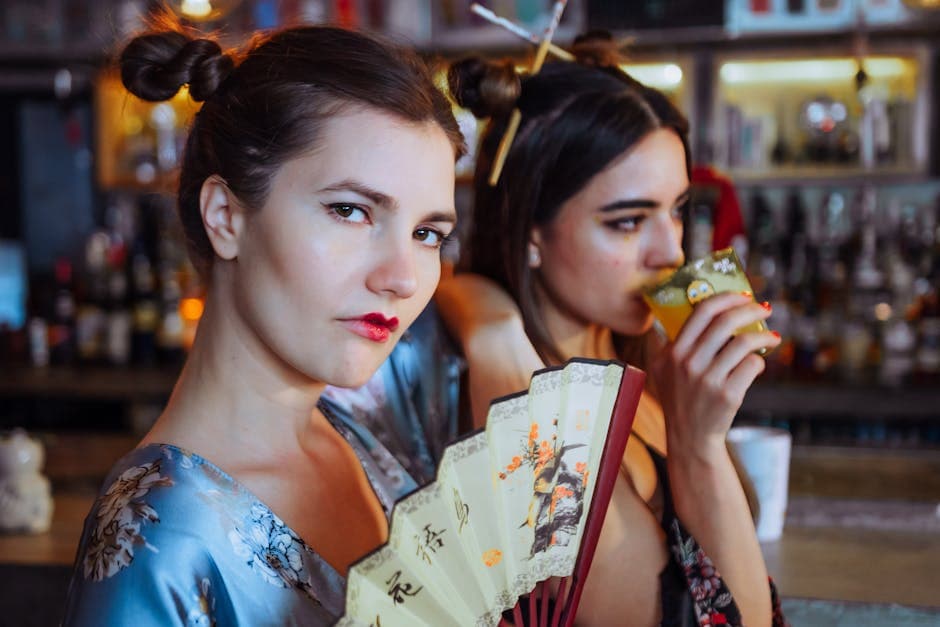 Two women in Asian-inspired attire posing with a hand fan, enjoying drinks in a bar.