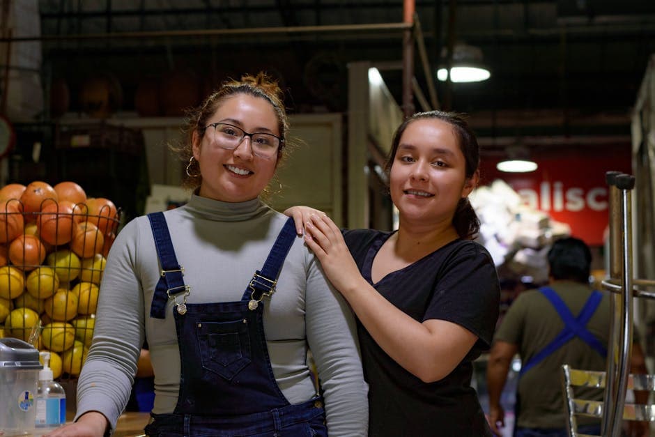 Two women smiling and posing at a fruit market in Ciudad de México, showcasing friendship and commerce.