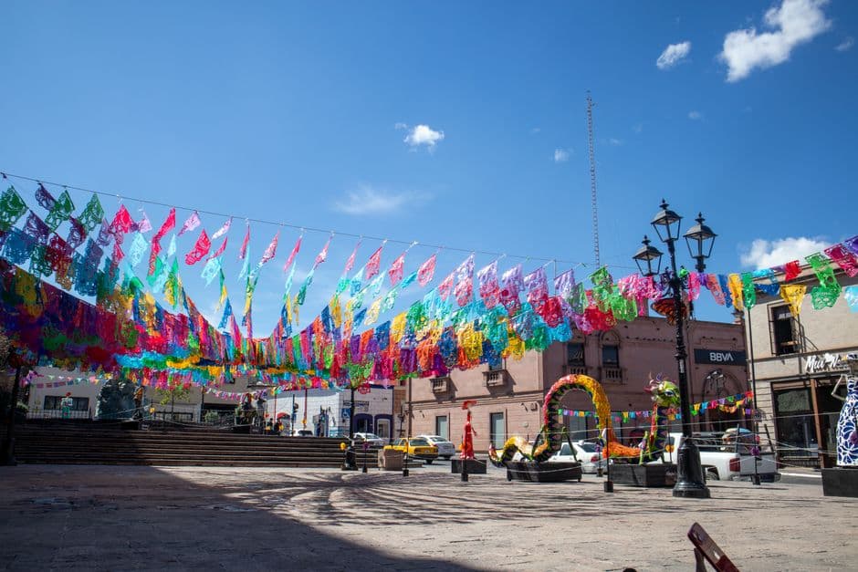 Colorful papel picado adorns a plaza in Saltillo, Mexico, under a clear blue sky during a festive celebration.