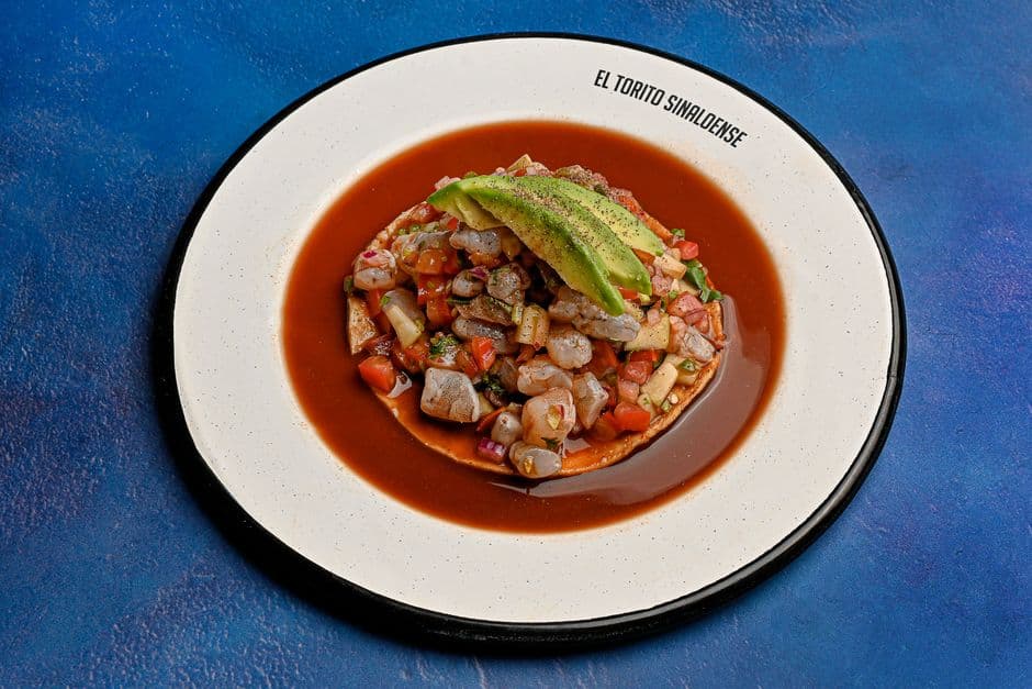 Colorful seafood tostada with avocado on a plate with blue backdrop.