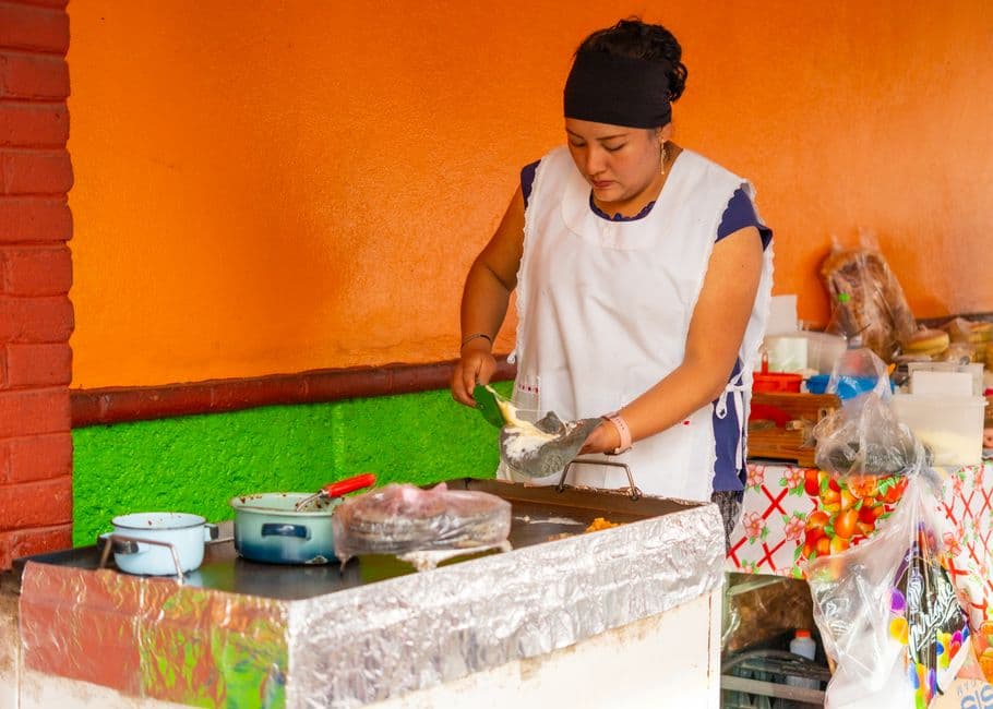 A Mexican woman skillfully prepares traditional street food indoors. Vibrant colors set the scene.
