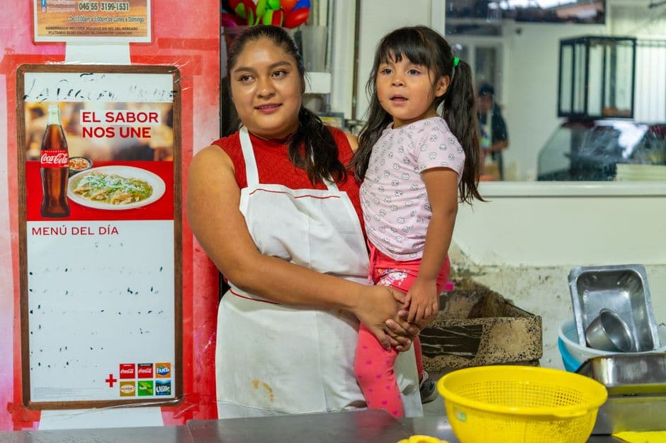 A mother holding her daughter in a local eatery near a 'Menu del Dia' sign.