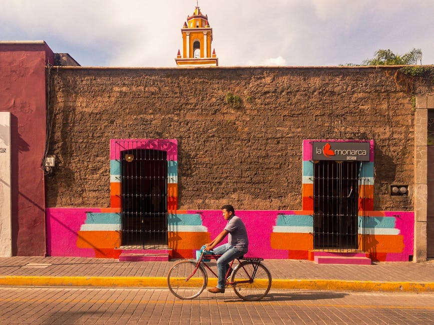 A man rides a bike past colorful architecture and a church tower in a Mexican city.