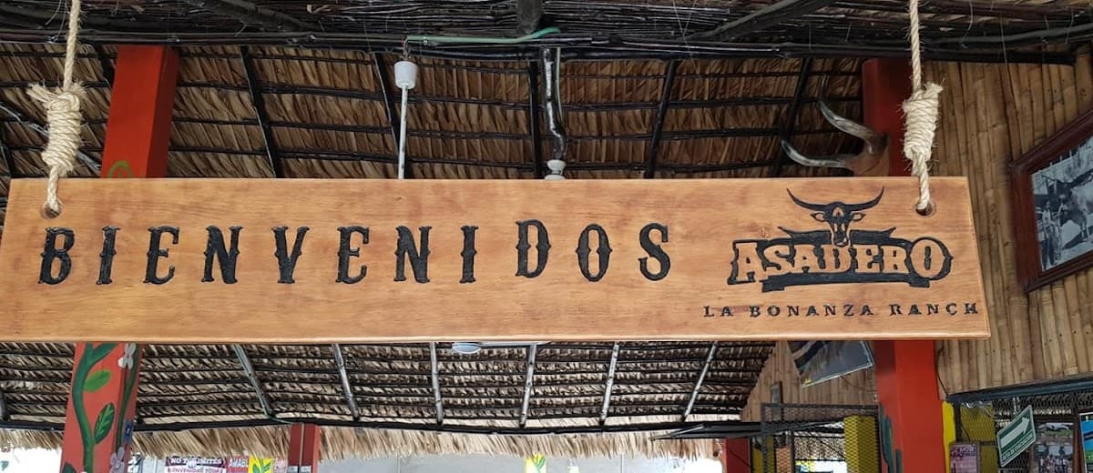 Exterior de La Bonanza Ranch Asadero con su señal en la carretera costera, mostrando la parrilla al aire libre y mesas bajo sombra