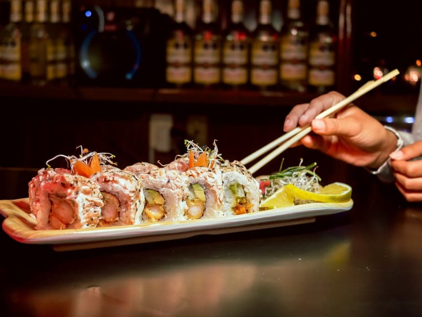 Close-up of gourmet sushi rolls with chopsticks in a cozy bar setting, highlighting fresh ingredients.