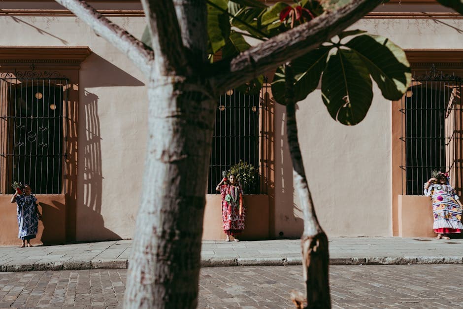 Women in traditional attire standing on a Oaxaca sidewalk, casting shadows in sunlight.