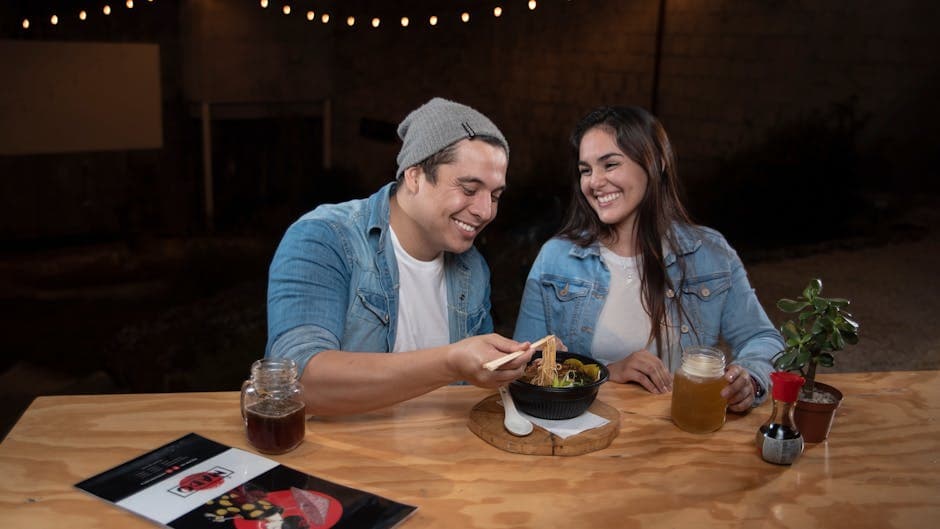 A couple enjoys ramen and drinks outdoors at night, sharing smiles and conversation.