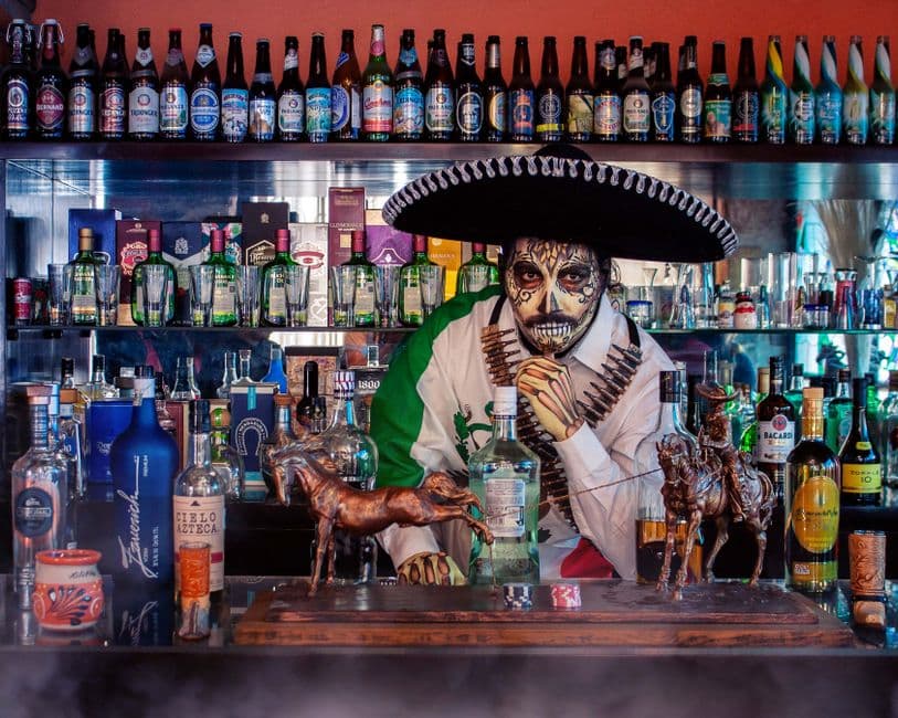 Vibrant bar scene with bartender dressed in Day of the Dead attire surrounded by bottles.