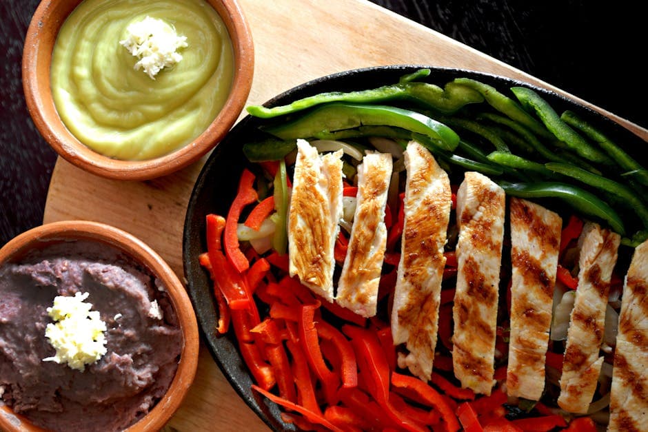 Sizzling grilled chicken fajitas with bell peppers, guacamole, and refried beans on a wooden board.
