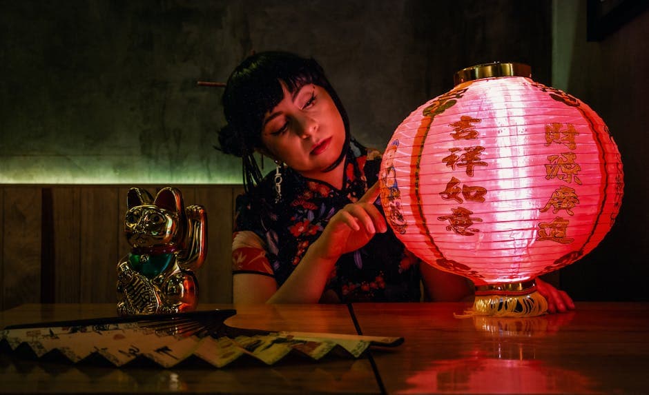 A woman in traditional attire admiring a red lantern with ornate designs beside a Maneki Neko in a dimly lit room.