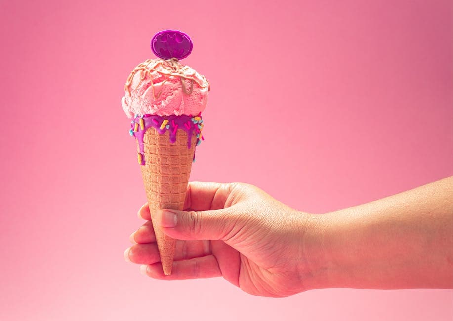 Pink ice cream cone with sprinkles and waffle cone held by a hand against a pink background in a studio setting.