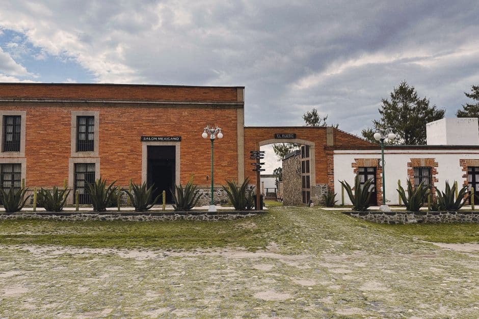 Red brick and white stucco Mexican building with cactus garden under a cloudy sky.