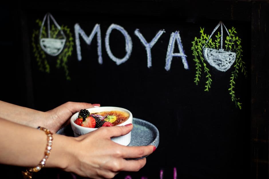 A person holding a crème brûlée dessert in front of a decorative chalkboard sign.