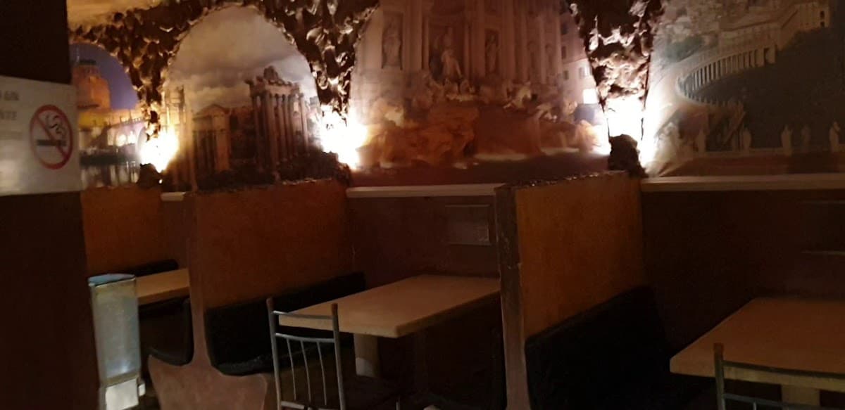 Exterior de Antica Roma con su fachada iluminada y mesas al aire libre, mostrando una botella de vino y platos de pasta en primer plano