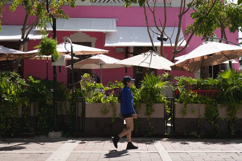 A woman walks past a vibrant pink cafe surrounded by lush greenery and umbrellas in sunny Merida.