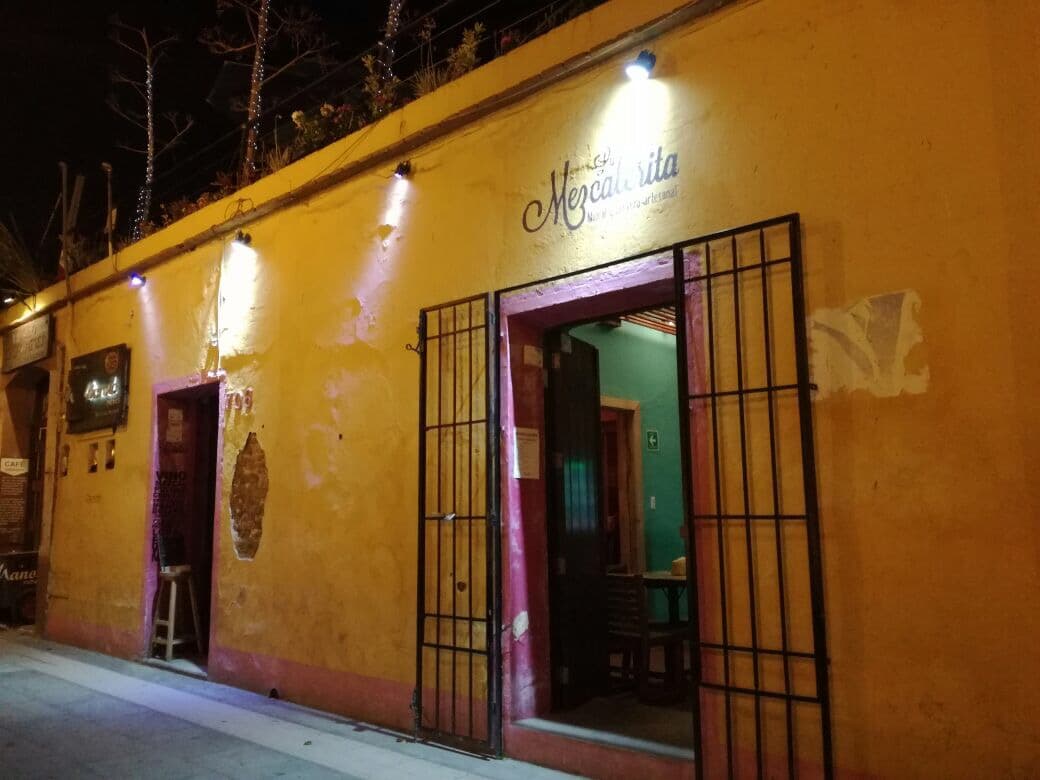 Exterior colorido de La Mezcalerita con su letrero de neón y una tabla de tlayudas recién servidas, frente a la calle Palmeras