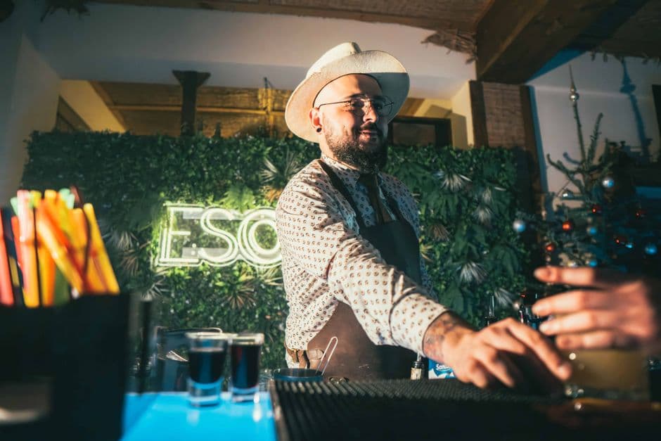 A stylish barman in a hat serves drinks at a lively bar with neon decor.