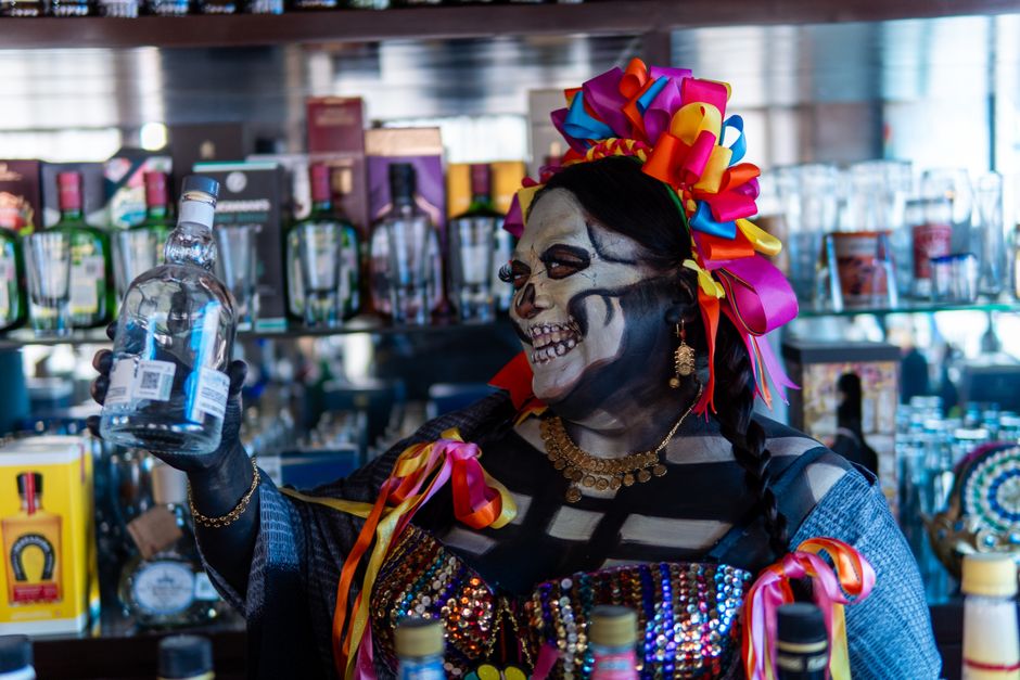 Colorful Catrina with traditional makeup holds a bottle in a festive Mexican bar.