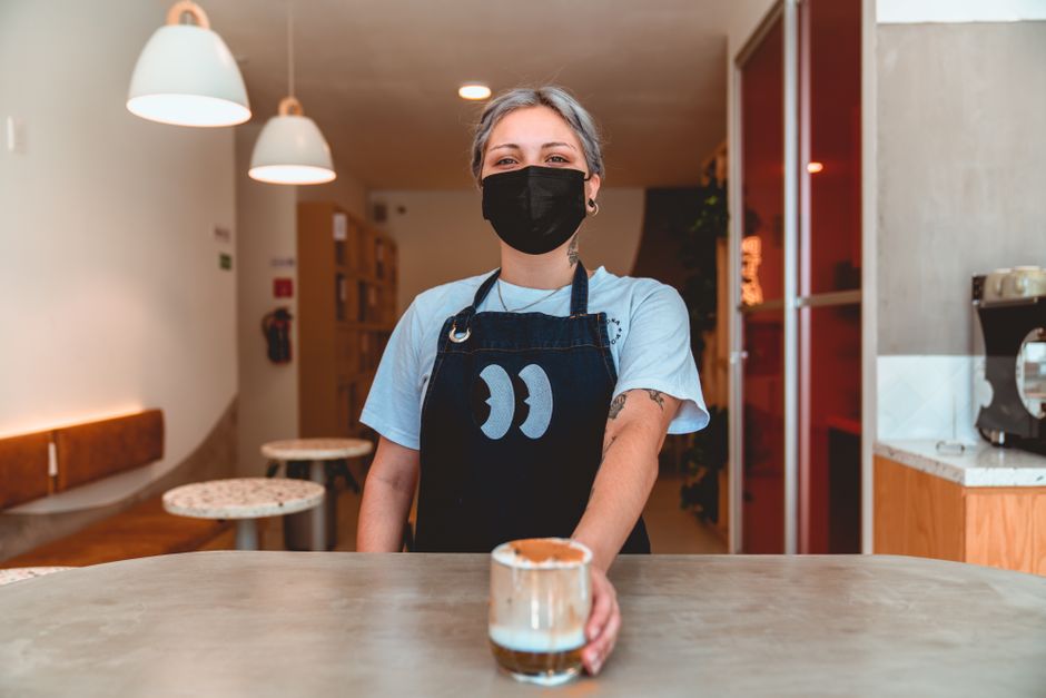A barista wearing a face mask serves coffee in a cozy café setting.