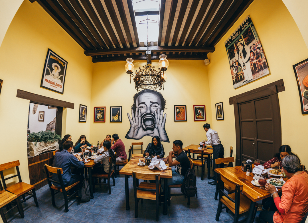 Interior del restaurante con mesas de madera, lámparas colgantes y el chef trabajando en la cocina abierta