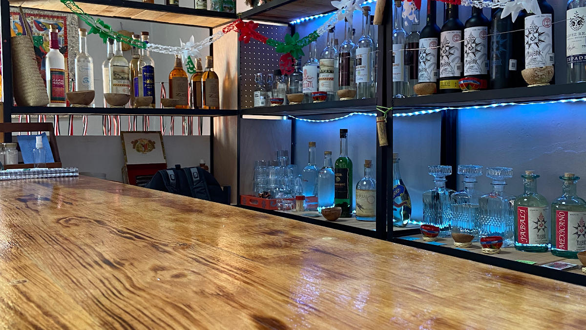 Bar interior mostrando botellas de mezcal alineadas y el bartender sirviendo una flight de tres mezcales