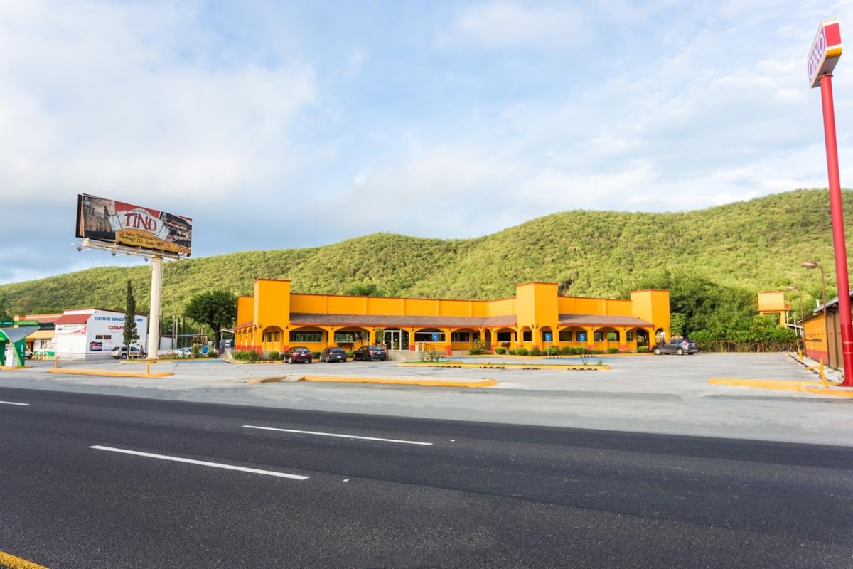 Exterior del Merendero Tino Santiago con su letrero rojo, una parrilla encendida y una mesa con tacos de carne asada