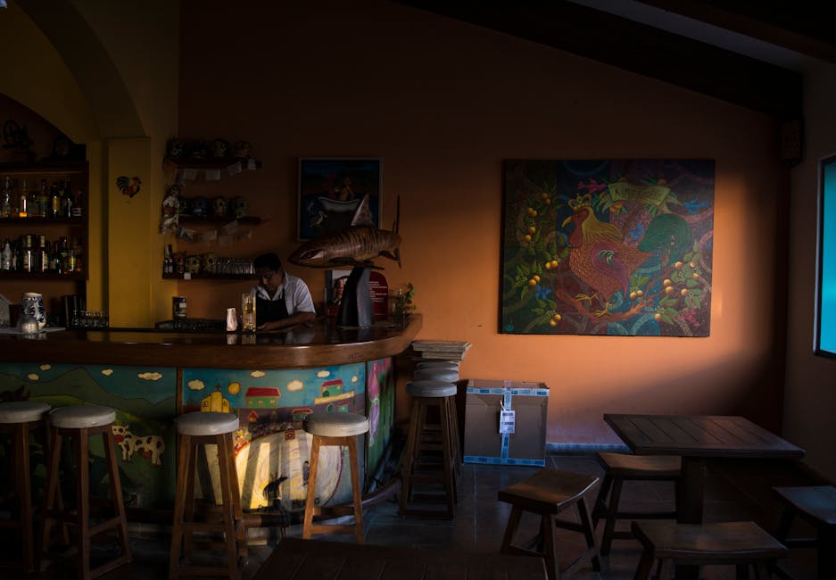 A warm bar interior featuring a colorful counter and stools, with a bartender working beneath vibrant artwork.